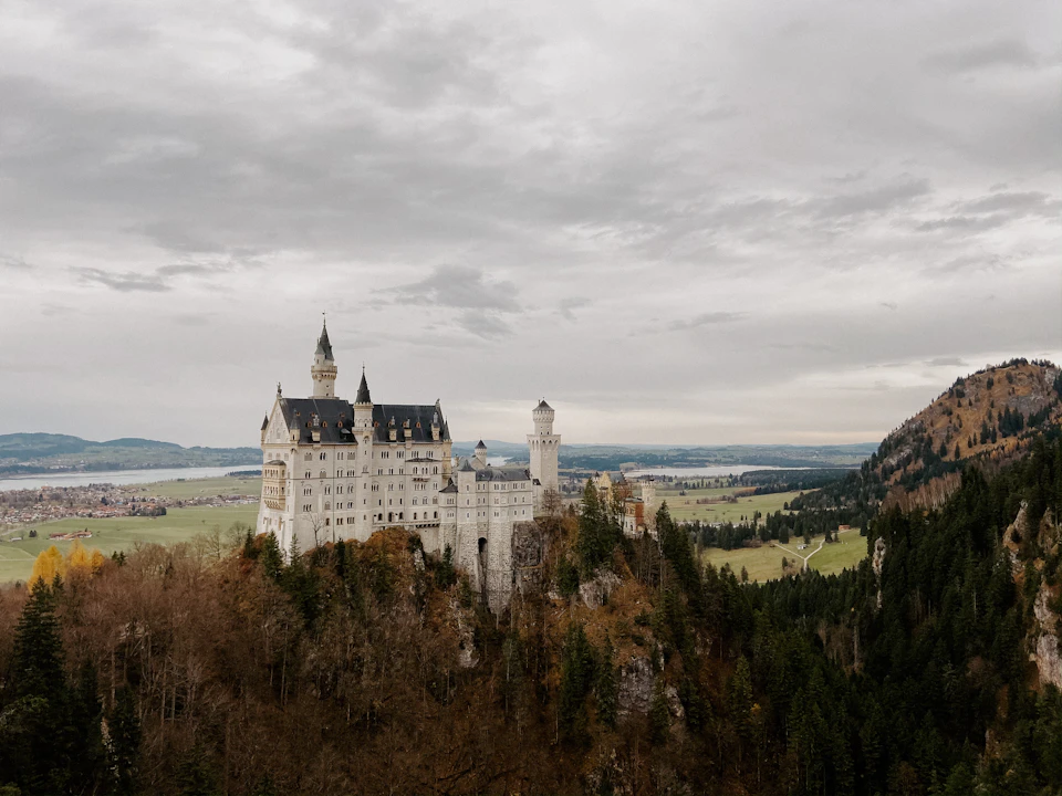 The image depicts a majestic castle perched on a cliff, overlooking a valley below. The castle is constructed from white stone and features multiple towers and turrets that reach towards the sky. The surrounding landscape includes trees and hills, creating a picturesque backdrop for the castle. The overcast sky casts a soft light over the scene, enhancing its grandeur.