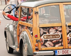 A vintage car with a wooden frame and a promotional decal featuring coffee beans and the words 'kafé o lait' is driven by a person in a red shirt. The side panel of the car displays contact information, and the rear window has a sticker reading 'Goodwood Motor Circuit'. The setting appears to be a street with spectators or bystanders visible in the background.