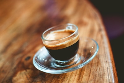 A clear glass cup filled with espresso sits on a matching glass saucer atop a wooden table. The rich, dark coffee is topped with a layer of creamy, light-colored crema. The background is blurred, putting focus on the coffee.