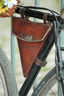 A vintage leather triangular bag is attached to the frame of a black bicycle. The bag is secured with two leather straps and features a buckle closure. The bicycle has a classic frame design, and part of the spoked wheel with a tire is visible. A basket filled with daisies is partially seen in the upper left corner.
