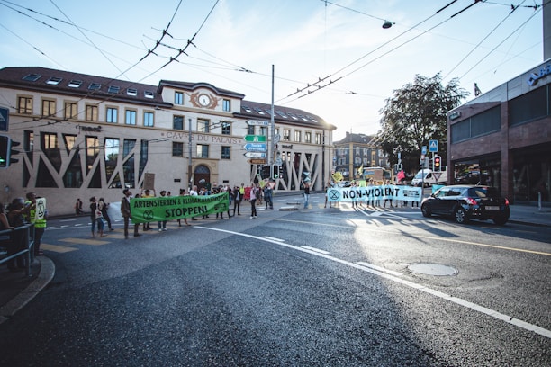 A group of protesters is gathered on a city street holding large banners. The banners contain messages about environmental issues and non-violence. The scene is set in an urban area with buildings, signs, and vehicles visible. The sky is clear with some light from the sun illuminating the street.