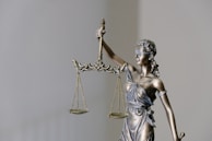 A bronze statue of Lady Justice holding scales in her right hand. She is blindfolded and wearing a classical Greco-Roman style robe. The background is plain and neutral colored.
