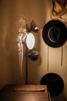 A room illuminated by a warm light features a burning incense stick on a wooden holder, with smoke curling upwards elegantly. A modern floor lamp casts a soft glow, highlighting the textured wall behind. Two hats are hung on the wall, adding a touch of personal decor.