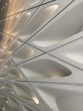A close-up view of a modern architectural surface featuring a pattern of curved, organic shapes. The surface is composed of smooth, white panels with recessed indentations, creating a wave-like effect. Soft ambient lighting highlights the texture and depth of the design.