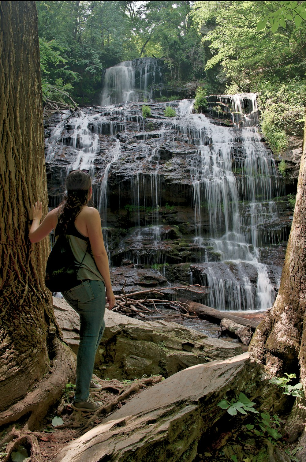 woman in black tank top and green pants standing on brown tree log near waterfalls during