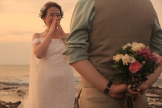 A bride in a white lace dress stands on a beach, covering her mouth with her hands in apparent surprise or joy. The groom is seen from the back, wearing a vest and shirt, holding a bouquet of colorful flowers behind his back. The scene is set during a sunset, providing a warm, romantic atmosphere.