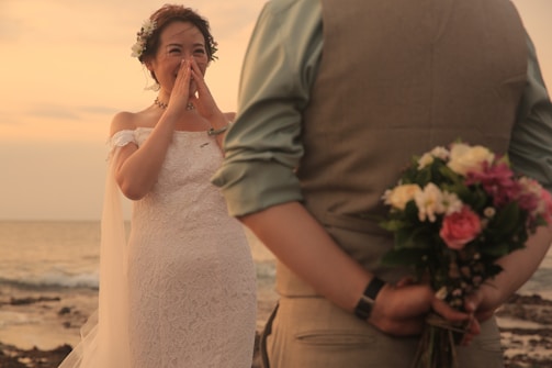 A bride in a white lace dress stands on a beach, covering her mouth with her hands in apparent surprise or joy. The groom is seen from the back, wearing a vest and shirt, holding a bouquet of colorful flowers behind his back. The scene is set during a sunset, providing a warm, romantic atmosphere.