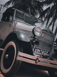 A vintage car with a black exterior and white-rimmed tires is parked on a brick road. The car bears a badge and is situated against a backdrop of tall palm trees. The angle of the photo emphasizes the car's front, including its headlights and the word 'VINTAGE' on the grill.