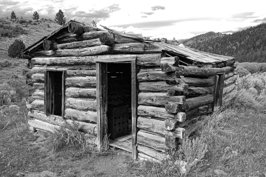 A rustic, weathered log cabin stands in a rural landscape, surrounded by rolling hills and sparse vegetation. The cabin appears aged with rough-hewn logs and an unfinished wooden door. In the background, distant hills and scattered trees create a serene, yet isolated atmosphere, under a partly cloudy sky.
