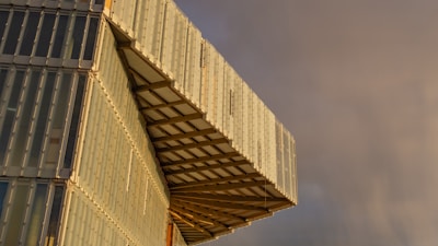 A modern architectural structure featuring an overhanging section composed of light-colored, translucent panels. The design includes a grid pattern and metallic framework. The background shows a cloudy sky with a mix of darker and lighter hues.