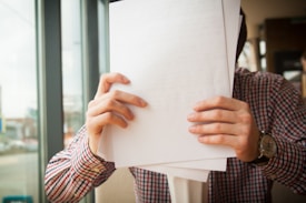 A person wearing a checkered shirt holds several sheets of paper in front of their face. Sunlight streams through a large window on the left, and a wristwatch is visible on one wrist.