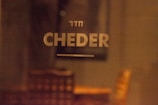 The image features the word 'CHEDER' in large, bold letters on a blurry background. Above it is a smaller text written in Hebrew. The setting appears to be indoors with warm lighting, and there’s a vague outline of furniture, such as chairs, in the background.