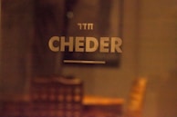 The image features the word 'CHEDER' in large, bold letters on a blurry background. Above it is a smaller text written in Hebrew. The setting appears to be indoors with warm lighting, and there’s a vague outline of furniture, such as chairs, in the background.