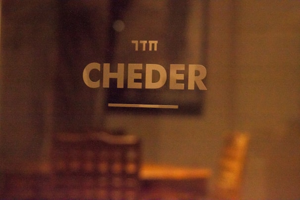 The image features the word 'CHEDER' in large, bold letters on a blurry background. Above it is a smaller text written in Hebrew. The setting appears to be indoors with warm lighting, and there’s a vague outline of furniture, such as chairs, in the background.