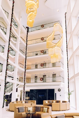 A spacious and modern hotel lobby with high ceilings and several floors visible above. The area is adorned with festive decorations, including Christmas wreaths and garlands. A large, elegant chandelier hangs prominently in the center. Comfortable seating with tan leather couches and cushions are arranged around a small coffee table.