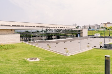 The foreground features a neatly manicured green lawn with a slight incline leading up to a modern building. The building is identified as the Kigali Convention Centre, with a sleek, glass-fronted entrance. The area in front of the entrance appears to be a spacious concrete plaza with a few planted shrubs. Surrounding the structure are additional landscaped areas and distant residential buildings.