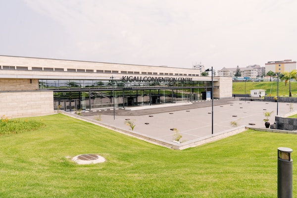 The foreground features a neatly manicured green lawn with a slight incline leading up to a modern building. The building is identified as the Kigali Convention Centre, with a sleek, glass-fronted entrance. The area in front of the entrance appears to be a spacious concrete plaza with a few planted shrubs. Surrounding the structure are additional landscaped areas and distant residential buildings.