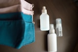 A set of face masks in teal and beige colors lies on a wooden surface, alongside three white spray bottles and a small transparent spray bottle.