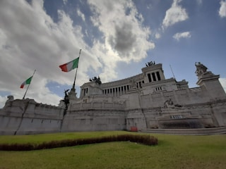 A grand, white marble monument with intricate sculptures and statues. Two tall flagpoles display the Italian flag, waving against a backdrop of partly cloudy skies. The structure features prominent columns and steps leading upwards, conveying a sense of historical grandeur.