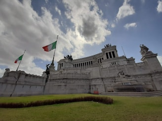 A grand, white marble monument with intricate sculptures and statues. Two tall flagpoles display the Italian flag, waving against a backdrop of partly cloudy skies. The structure features prominent columns and steps leading upwards, conveying a sense of historical grandeur.