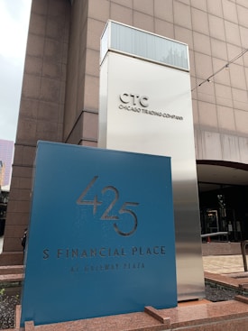 A modern building exterior with signage for the Chicago Trading Company and 425 S Financial Place at Gateway Plaza. The structure features a reflective metallic sign tower with bold lettering. The area appears to be part of a commercial or financial district.