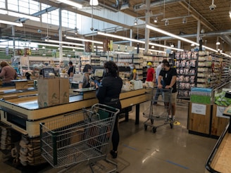 A busy supermarket scene with several people at the checkout counters. Customers are waiting in line with shopping carts filled with groceries. The store has various sections with shelves stocked with products. The ambiance is well lit with large overhead lights and natural light coming through the large windows.