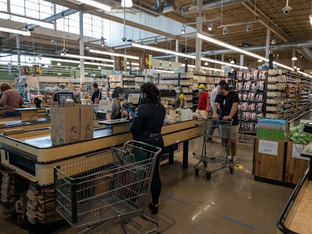 A busy supermarket scene with several people at the checkout counters. Customers are waiting in line with shopping carts filled with groceries. The store has various sections with shelves stocked with products. The ambiance is well lit with large overhead lights and natural light coming through the large windows.