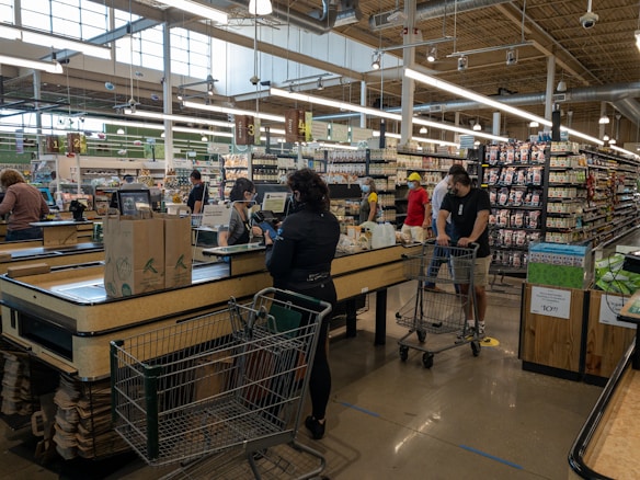 A busy supermarket scene with several people at the checkout counters. Customers are waiting in line with shopping carts filled with groceries. The store has various sections with shelves stocked with products. The ambiance is well lit with large overhead lights and natural light coming through the large windows.