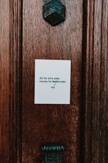 A close-up of a textured wooden surface with a small white paper attached to it. The paper contains a printed phrase that reads, 'Art for art's sake / money for God’s sake / my'. Above the paper, there is a dark metallic geometric object that resembles a decorative fixture.