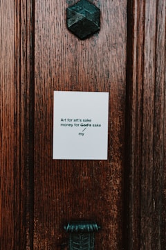 A close-up of a textured wooden surface with a small white paper attached to it. The paper contains a printed phrase that reads, 'Art for art's sake / money for God’s sake / my'. Above the paper, there is a dark metallic geometric object that resembles a decorative fixture.