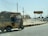 A man is driving a yellow and green auto-rickshaw on a sunlit road. To the right, there is a signboard with the words 'Karachi The City of Lights'. The surrounding area looks dry and somewhat deserted, with a few more vehicles on the road and power lines overhead.