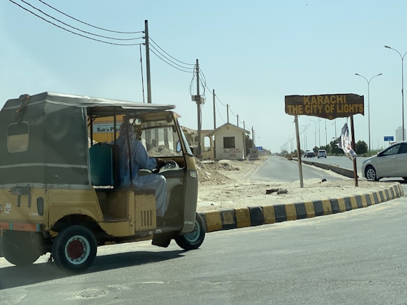 A man is driving a yellow and green auto-rickshaw on a sunlit road. To the right, there is a signboard with the words 'Karachi The City of Lights'. The surrounding area looks dry and somewhat deserted, with a few more vehicles on the road and power lines overhead.