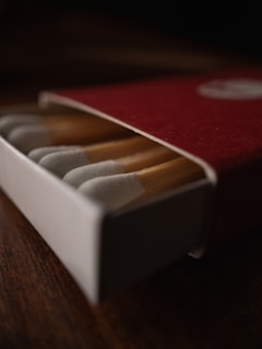 A close-up of a partially opened matchbox with several wooden matches inside, placed on a dark wooden surface. The focus is sharp on the tips of the matches.