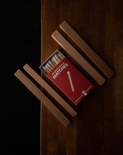 A red box of wooden matches is partially open, revealing a row of matchsticks inside. It is placed on a wooden surface with several wooden sticks arranged parallel and perpendicular to the box.