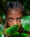 A person with braided hair is partially obscured by large green leaves. Their face is visible, with an intense gaze directed towards the viewer. The lighting highlights their complexion and creates a natural, outdoor setting.