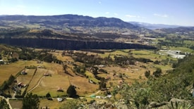 A vast landscape with a mixture of open fields and scattered houses, surrounded by mountains in the distance. The land is divided into rectangular plots, with various shades of green and brown. A deep ravine cuts across the center, adding dramatic depth to the scenery. Sparse trees are dotted around the fields, and the sky is clear with minimal clouds.