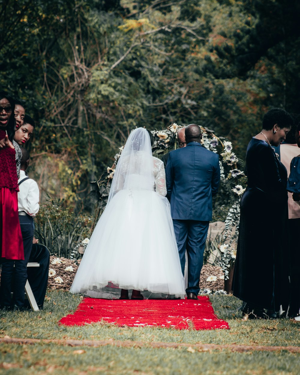 Braut und Bräutigam stehen tagsüber auf roten Textilien, umgeben von Bäumen