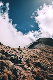 A rugged mountain trail with hikers making their way up a steep slope. The foreground is filled with rocky terrain and dry, golden grasses, while the background features a dramatic sky with clouds partially covering the mountain peak, adding a sense of mystery and adventure.