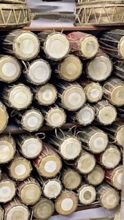 A collection of traditional drums is neatly arranged on shelves, showcasing the circular drumheads and intricate rope lacing. The drums vary in color, with natural wood and darker tones highlighted.