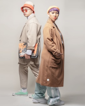 Two individuals are wearing fashionable, oversized coats in neutral tones. One person is in a beige coat, beige bucket hat, orange sunglasses, and green sneakers, holding a folder with an abstract print. The other person is dressed in a light brown coat, yellow beanie, purple hairstyle, and white sneakers, featuring a confident expression.
