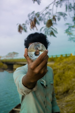 A person holding a glass sphere in front of their face, creating a lens-like effect that inverts and distorts the background. The setting features an outdoor environment with a bridge, river, and overhanging tree branches.