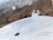 A metal topographical indicator displaying various landmarks, distances, and heights is prominently in focus. The background reveals an out-of-focus rocky terrain and a blurred figure of a person, suggesting an outdoor, elevated setting.
