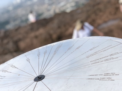 A metal topographical indicator displaying various landmarks, distances, and heights is prominently in focus. The background reveals an out-of-focus rocky terrain and a blurred figure of a person, suggesting an outdoor, elevated setting.