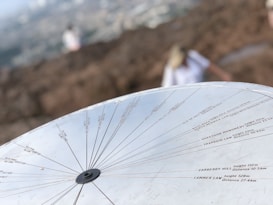 A metal topographical indicator displaying various landmarks, distances, and heights is prominently in focus. The background reveals an out-of-focus rocky terrain and a blurred figure of a person, suggesting an outdoor, elevated setting.