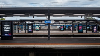 A train station with multiple platforms, advertising signs displaying messages related to drug tests, and a sign indicating Richmond station. Overcast skies are visible above the platform roof.