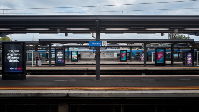 A train station with multiple platforms, advertising signs displaying messages related to drug tests, and a sign indicating Richmond station. Overcast skies are visible above the platform roof.