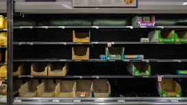 Several supermarket shelves with mostly empty cardboard boxes, a few cartons of eggs left. The surrounding area is dimly lit with a focus on the shelves displaying mostly empty spaces where products should be placed. Labels and price tags are visible below the shelves.