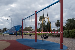 A deserted playground with swings and climbing structures is cordoned off with red and white tape. The sky is overcast, and trees and park benches are visible in the background. The ground is covered with gravel and rubber safety mats.