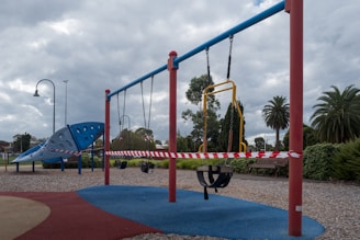 A deserted playground with swings and climbing structures is cordoned off with red and white tape. The sky is overcast, and trees and park benches are visible in the background. The ground is covered with gravel and rubber safety mats.