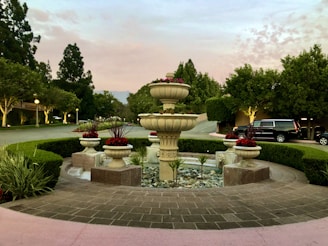 A beautifully manicured garden setting features a prominent stone fountain surrounded by brick paving and circular hedges. The fountain is adorned with potted red flowers, and a driveway in the background is lined with trees and bushes under a soft pink and blue evening sky.